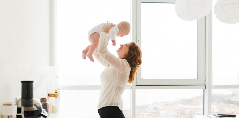 Portrait of beautiful happy woman standing and holding her pretty little baby in hands with big windows on background